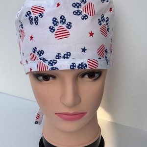 May include: A white surgical scrub cap featuring a pattern of red and blue paw prints and stars. The paw prints have a striped design, and the stars are red. The cap is tied at the back and is displayed on a mannequin head.