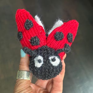 Peut inclure: Jouet coccinelle tricoté à la main. La coccinelle a des ailes rouges avec des pois noirs, une tête noire et des yeux blancs. Les ailes sont légèrement ouvertes et le jouet est en fil doux.