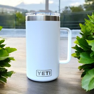 K&ouml;nnte beinhalten: Wei&szlig;er Yeti Rambler Becher mit schwarzem Griff und dem Yeti-Logo auf der Vorderseite.