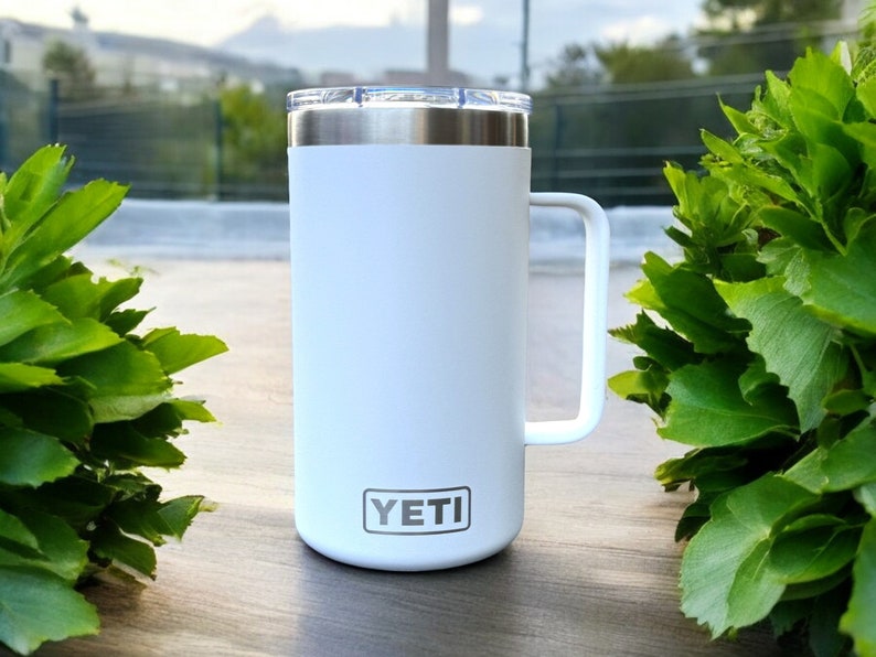 K&ouml;nnte beinhalten: Wei&szlig;er Yeti Rambler Becher mit schwarzem Griff und dem Yeti-Logo auf der Vorderseite.