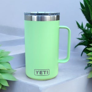 K&ouml;nnte beinhalten: Eine hellgr&uuml;ne YETI-Reisebecher mit einem Edelstahldeckel und einem Griff. Der Becher steht auf einer wei&szlig;en Oberfl&auml;che.
