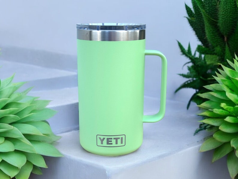 K&ouml;nnte beinhalten: Eine hellgr&uuml;ne YETI-Reisebecher mit einem Edelstahldeckel und einem Griff. Der Becher steht auf einer wei&szlig;en Oberfl&auml;che.
