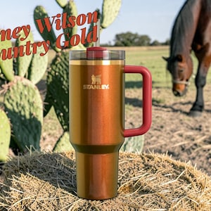 Puede incluir: Un vaso Stanley de color cobre con asa y tapa burdeos, colocado sobre una paca de heno. El vaso tiene el logotipo de Stanley y el texto "Lainey Wilson Country Gold". Un caballo pasta al fondo.