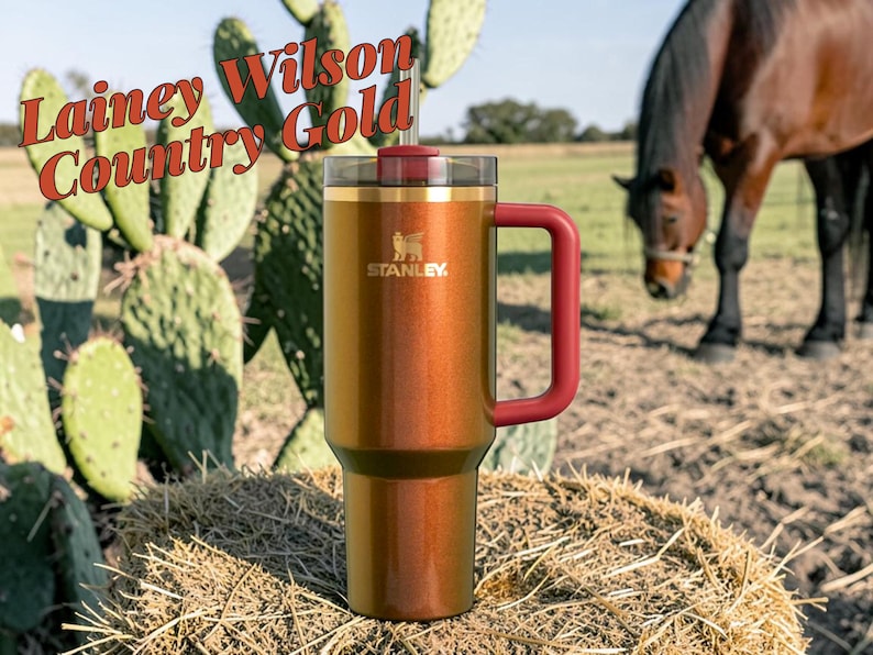 Puede incluir: Un vaso Stanley de color cobre con asa y tapa burdeos, colocado sobre una paca de heno. El vaso tiene el logotipo de Stanley y el texto "Lainey Wilson Country Gold". Un caballo pasta al fondo.