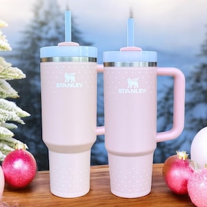 May include: Two pink stainless steel travel mugs with handles and lids. Both mugs have a white polka dot pattern and the Stanley logo. The mugs are sitting on a wooden surface with Christmas ornaments.