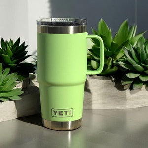 May include: A green YETI travel mug with a stainless steel lid and handle. The mug is sitting on a table with a white surface.