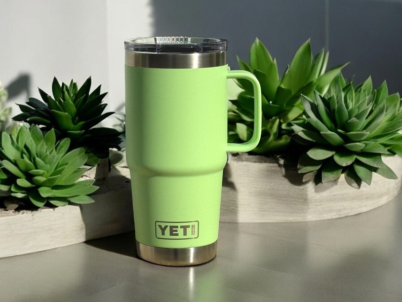 May include: A green YETI travel mug with a stainless steel lid and handle. The mug is sitting on a table with a white surface.