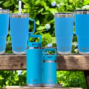 Op de afbeelding: Een set van vijf blauwe YETI-bekers, waaronder twee met handvatten, twee kleinere flessen en een met een rietje. De bekers staan allemaal op een houten bank met een groene gras achtergrond.