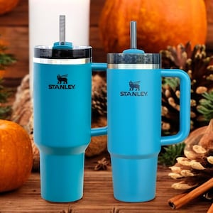 May include: Two blue stainless steel travel mugs with handles and lids. Both mugs have a black Stanley logo with a winged creature. The mugs are on a wooden surface with pumpkins and pine cones.