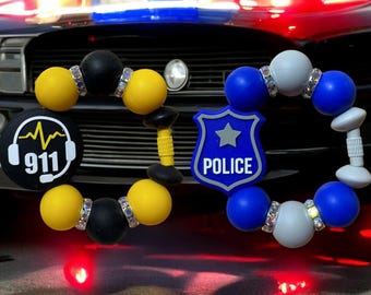 May include: Two silicone bead bracelets with charms. One bracelet is yellow and black with a 911 emergency call charm. The other bracelet is blue and white with a police badge charm.