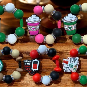 May include: Three colorful silicone bracelets with charms featuring coffee cup designs. The charms include a pink coffee cup with the word "MOM" on it, a green coffee cup with the words "TRIPS & SIPS" on it, and a brown coffee cup with the text "coffee is my blood type" on it.