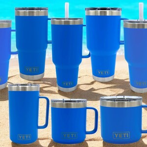 Op de afbeelding: Acht blauwe YETI bekers met deksels en handvatten. Sommige bekers hebben rietjes. De bekers staan op een zanderig oppervlak.