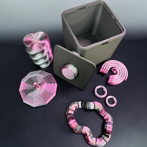 May include: An assortment of metallic objects in shades of pink, silver, and gray. The items include a square container with a lid, a stack of spiral-shaped pieces, an octagonal design, a curved shape, two rings, and a segmented bracelet.