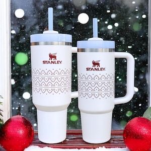 May include: Two white Stanley travel mugs with a red and white patterned design. The mugs have a red logo that says "STANLEY" and a red bear graphic. The mugs have lids with built-in straws.