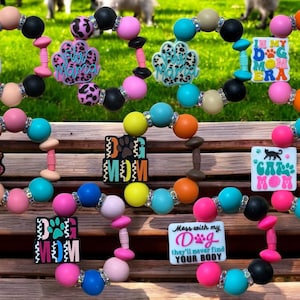 May include: Assortment of colorful beaded bracelets with various dog mom themed charms. The charms include "My Kids Have Paws", "Dog Mom", "Furry Mama", "In My Dog Mom Era", "Mess with my Dog they'll never find your body", and "Cat Mom".