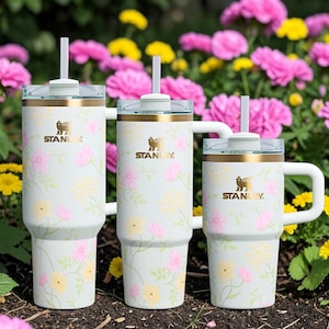 May include: Three Stanley tumblers with floral patterns. The tumblers are white with pink and yellow flowers, gold accents, and the Stanley logo. Each tumbler has a white straw and a clear lid. The background features pink and yellow flowers.