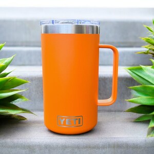 K&ouml;nnte beinhalten: Orangefarbener YETI Rambler 14 oz Becher mit Edelstahldeckel und -griff. Der Becher steht auf einer Betontreppe.