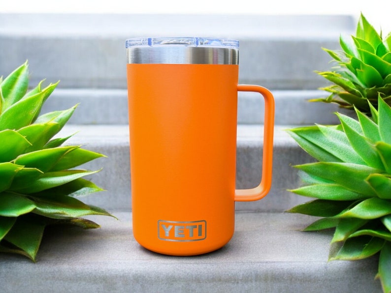 K&ouml;nnte beinhalten: Orangefarbener YETI Rambler 14 oz Becher mit Edelstahldeckel und -griff. Der Becher steht auf einer Betontreppe.