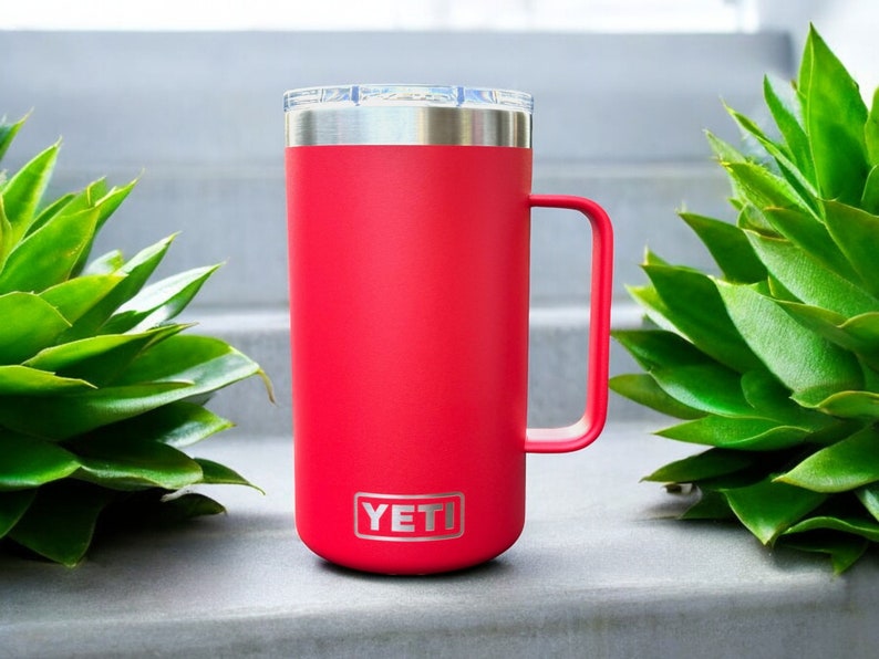 K&ouml;nnte beinhalten: Eine rote YETI Rambler 14 oz Tasse mit einem Edelstahldeckel und -griff. Die Tasse steht auf einer grauen Oberfl&auml;che.