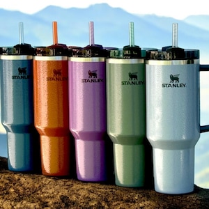 May include: Five stainless steel travel mugs with lids and straws. The mugs are in different colours: blue, orange, purple, green, and silver. The mugs have the Stanley logo on them.