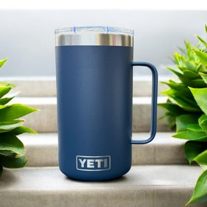 K&ouml;nnte beinhalten: Eine blaue YETI Rambler 14 oz Tasse mit einem Edelstahldeckel und -griff. Die Tasse steht auf einer Betontreppe.