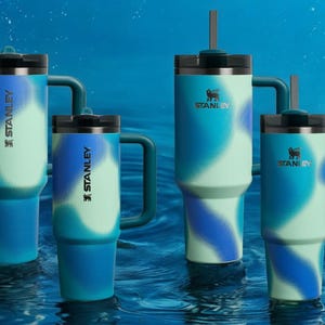 Könnte beinhalten: Vier Edelstahl-Reisebecher mit einem blauen und grünen Ombre-Design. Die Becher tragen das Stanley-Logo und einen schwarzen Griff. Die Becher schweben in einem Wasserbecken.