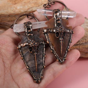 Puede incluir: Dos colgantes de punta de flecha con cristales de cuarzo. Los colgantes tienen un marco de metal color bronce y un diseño de calavera de pájaro. Las puntas de flecha son de piedra gris y marrón. Los colgantes están en un llavero.