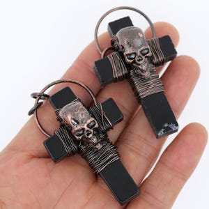Könnte beinhalten: Zwei schwarze Onyx-Kreuzanhänger mit Totenkopf-Design. Jedes Kreuz ist mit Kupferdraht umwickelt und hat einen detaillierten Totenkopf-Anhänger.
