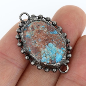 Op de afbeelding: Ovaalvormige hanger met een blauwe en bruine steen gezet in een zilverkleurig metalen frame. Het frame is versierd met een parelrand.