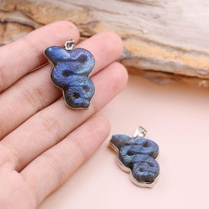 Puede incluir: Dos colgantes con forma de serpiente con borde plateado. Los colgantes son una mezcla de tonos azules y morados. El diseño de la serpiente es detallado, con un lazo en la parte superior para una cadena. El fondo es rosa claro.
