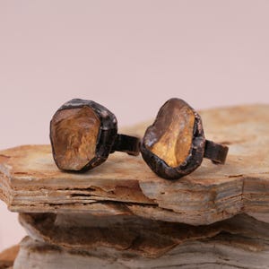 May include: Two rings with a rough, irregular, natural stone setting. The stones are a light amber color and are set in a dark brown metal.