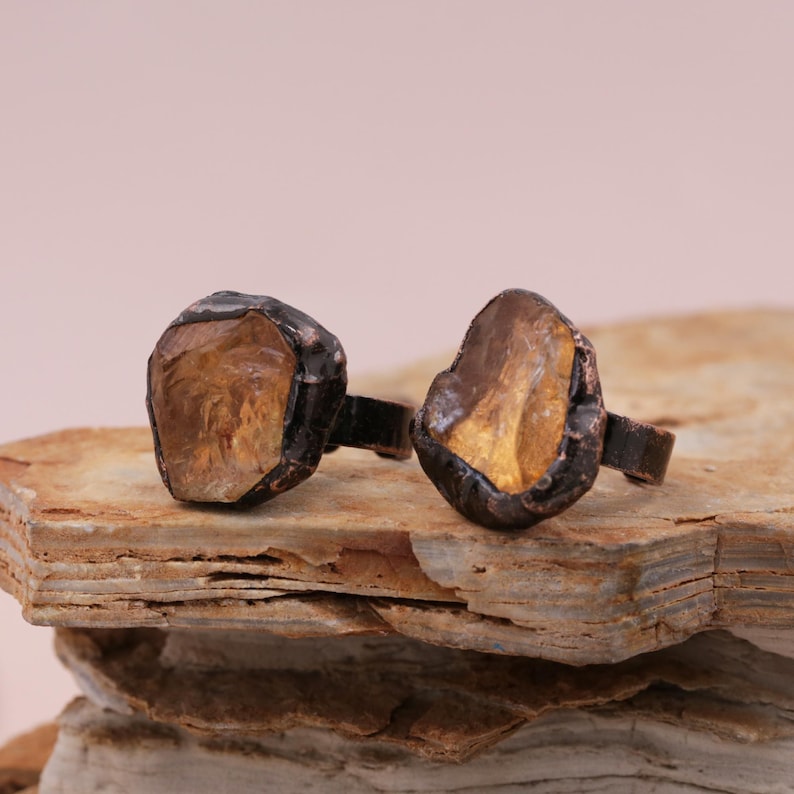 May include: Two rings with a rough, irregular, natural stone setting. The stones are a light amber color and are set in a dark brown metal.