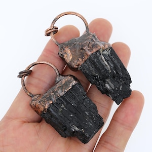 May include: Two black tourmaline crystal pendants with copper wire wraps. The crystals are raw and have a rough, natural texture. The copper wire is wrapped around the top of each crystal and forms a loop for hanging.