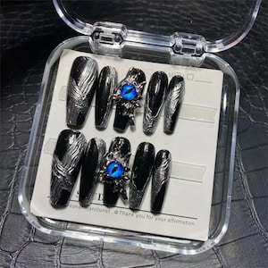 Puede incluir: Un juego de uñas postizas negras con detalles plateados y adornos de ojos azules. Las uñas se muestran en una caja de plástico transparente.