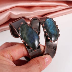 May include: A pair of copper-toned metal bracelets, each featuring a large, oval-shaped labradorite stone with blue and green iridescence. The stones are set in a decorative, beaded bezel. The bracelets have a curved, open design.
