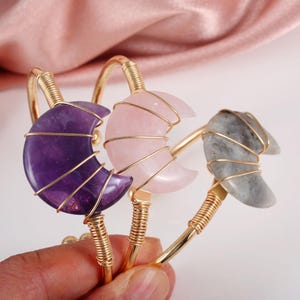Puede incluir: Tres pulseras doradas, cada una con una gema en forma de media luna. Las gemas son amatista, cuarzo rosa y una piedra gris. Las pulseras tienen un diseño con alambre alrededor de las gemas.
