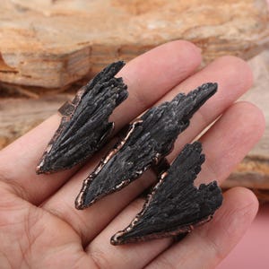 May include: Three black kyanite rings with a bronze-coloured metal band. The kyanite has a rough, textured appearance. The rings are displayed on a hand against a neutral background.