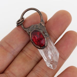 Op de afbeelding: Een hanger van helder kwartskristal met een rode edelsteen ingelegd in koperdraad. De hanger heeft een lus om aan een ketting te bevestigen.