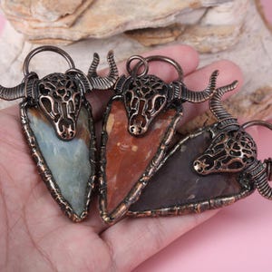 May include: Three handcrafted pendants with a bull's head design. Each pendant has a different coloured stone: blue-green, red-brown, and dark grey. The bull's head is made of a bronze-coloured metal with intricate cut-out details.