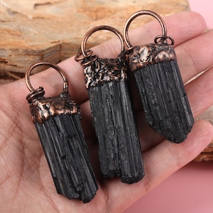 May include: Three black tourmaline pendants with copper-colored metal caps and loops. The raw, cylindrical crystals are a deep black, and the metal has a textured, aged appearance. The pendants are displayed against a neutral background.