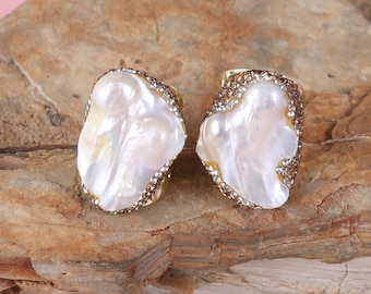 May include: Two gold-toned rings, each featuring a large, irregular white pearl. The pearls are surrounded by a band of small, sparkling crystals. The rings are displayed on a textured, brown surface.