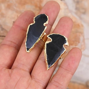 May include: Two black arrowhead shaped rings with gold trim. The rings are on a person's fingers.