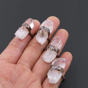 Op de afbeelding: Vier ringen met heldere kwartskristallen gezet in koperen banden. De ringen worden gedragen op een hand, waarbij elke vinger een andere ring toont.