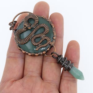 以下が含まれることがあります： 蛇のデザインが施された緑色の石が付いた銅製のペンダント。ペンダントは、緑色の石のビーズがついた銅製のチェーンに繋がっています。