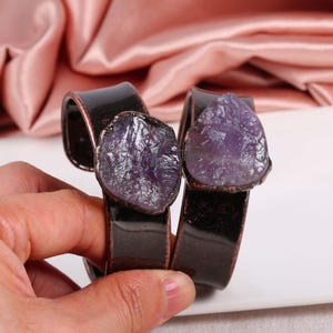 May include: Two dark brown metal bracelets, each featuring a raw, purple amethyst stone. The bracelets have a glossy finish and a rustic, natural aesthetic. The amethyst stones are irregularly shaped and add a unique touch.