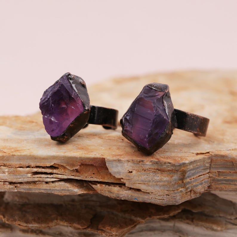 May include: Two rings with purple amethyst stones set in copper. The rings are on a rough, brown, layered rock surface.