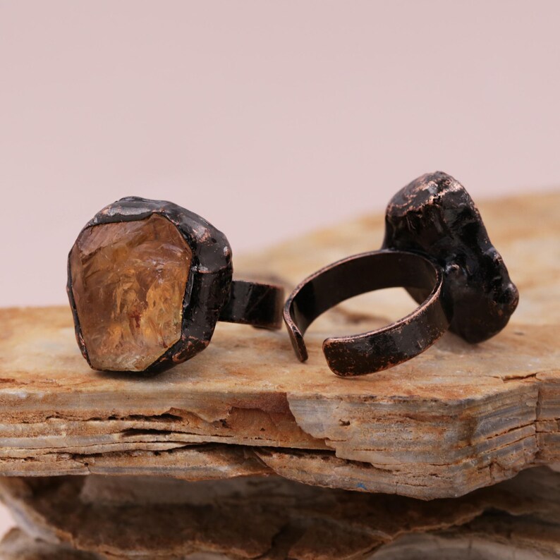 May include: Two adjustable rings with a copper finish. Each ring features a large, irregular shaped, clear, yellow-brown gemstone.