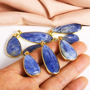 May include: A set of six blue kyanite gemstone pendants with gold-plated settings. The pendants are in the shape of teardrops and are arranged on a hand.