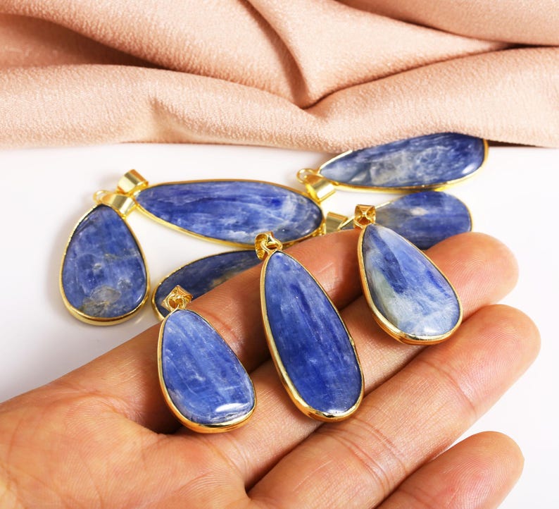 May include: A set of six blue kyanite gemstone pendants with gold-plated settings. The pendants are in the shape of teardrops and are arranged on a hand.