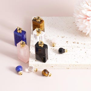May include: Assortment of small, gemstone bottles with gold caps. The bottles are made of various stones, including tiger's eye, clear quartz, lapis lazuli, rose quartz, and black obsidian. The bottles are arranged on a white marble surface.